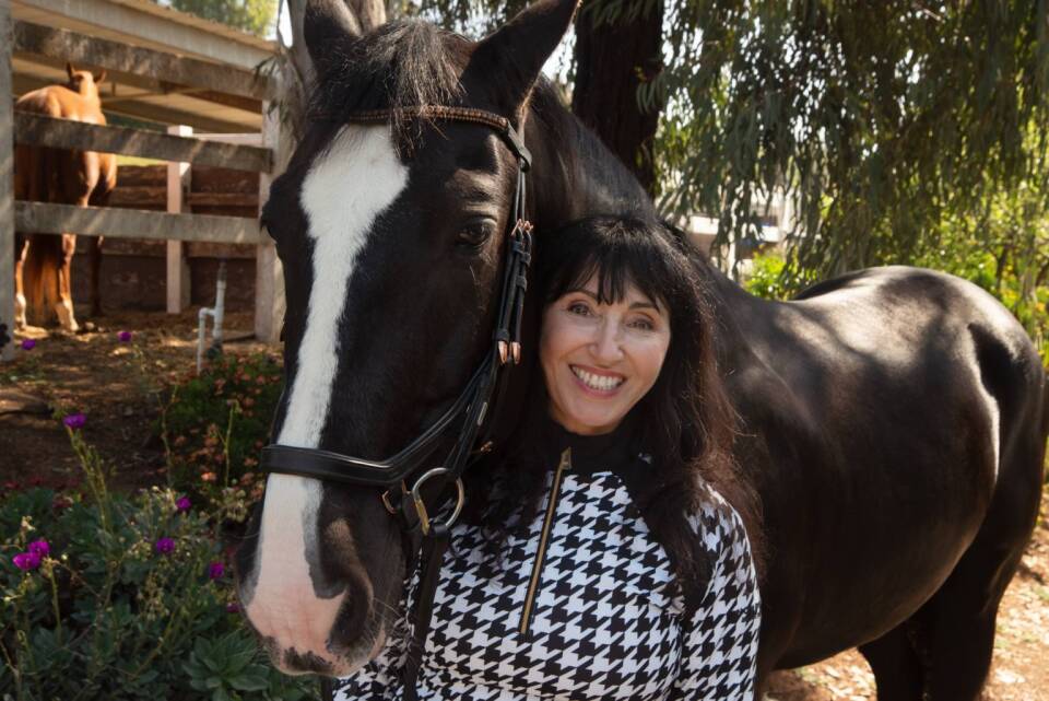 A woman with long brown hair wearing a black and white printed jacket stands smiling next to a black and white horse