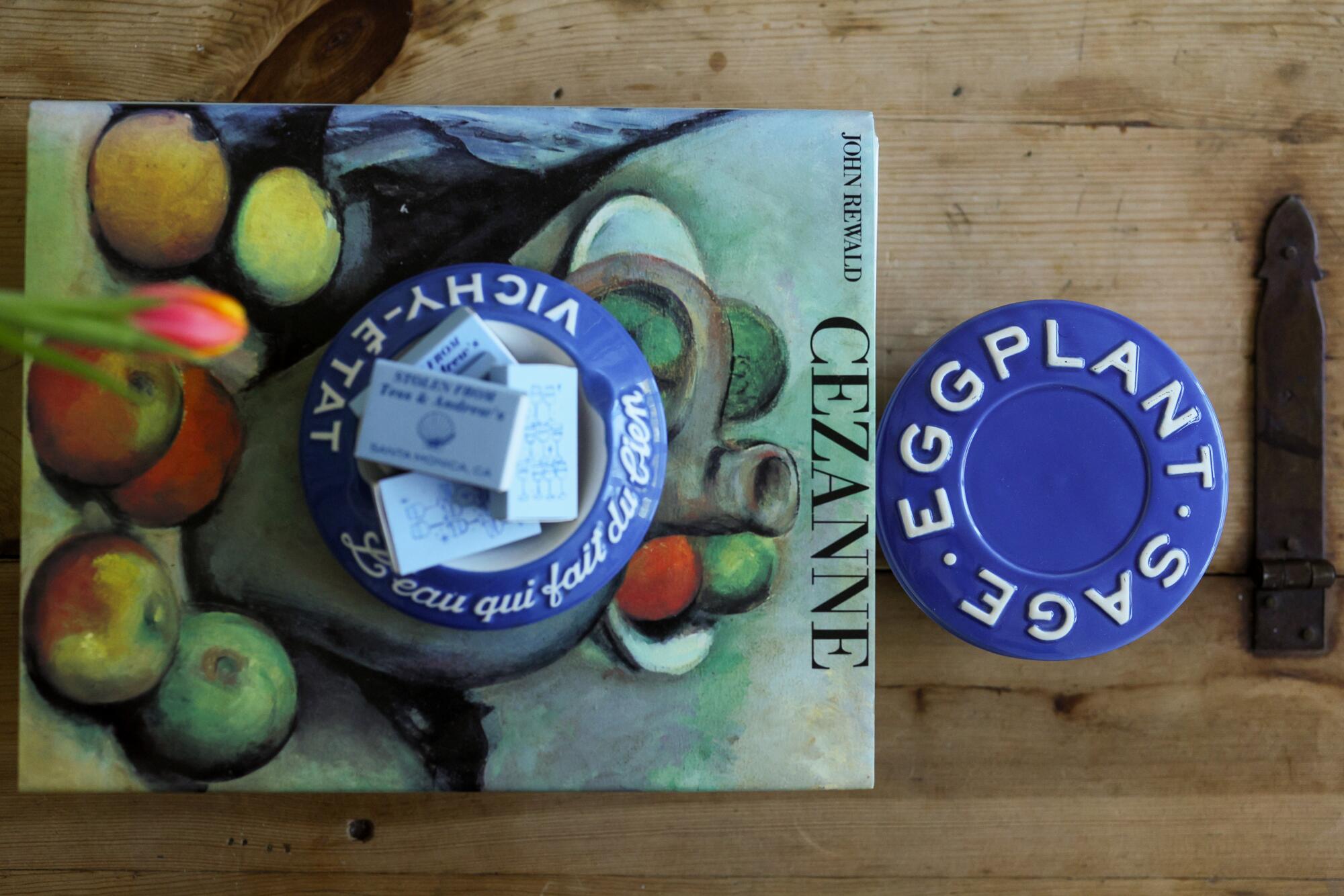 A blue vintage ashtray rests on top of a Cezanne book next to a vintage ceramic container on top a chest.