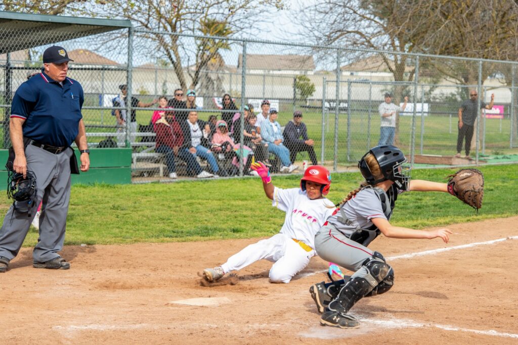 Young girl slides to homebase as umpire watches from side and catcher squats in anticipation of a throw.