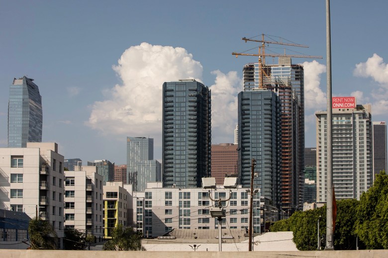 Mid-rise apartment buildings sit in the foreground as glass high-rises and two construction cranes rise behind them against a blue sky with large white clouds in downtown Los Angeles.