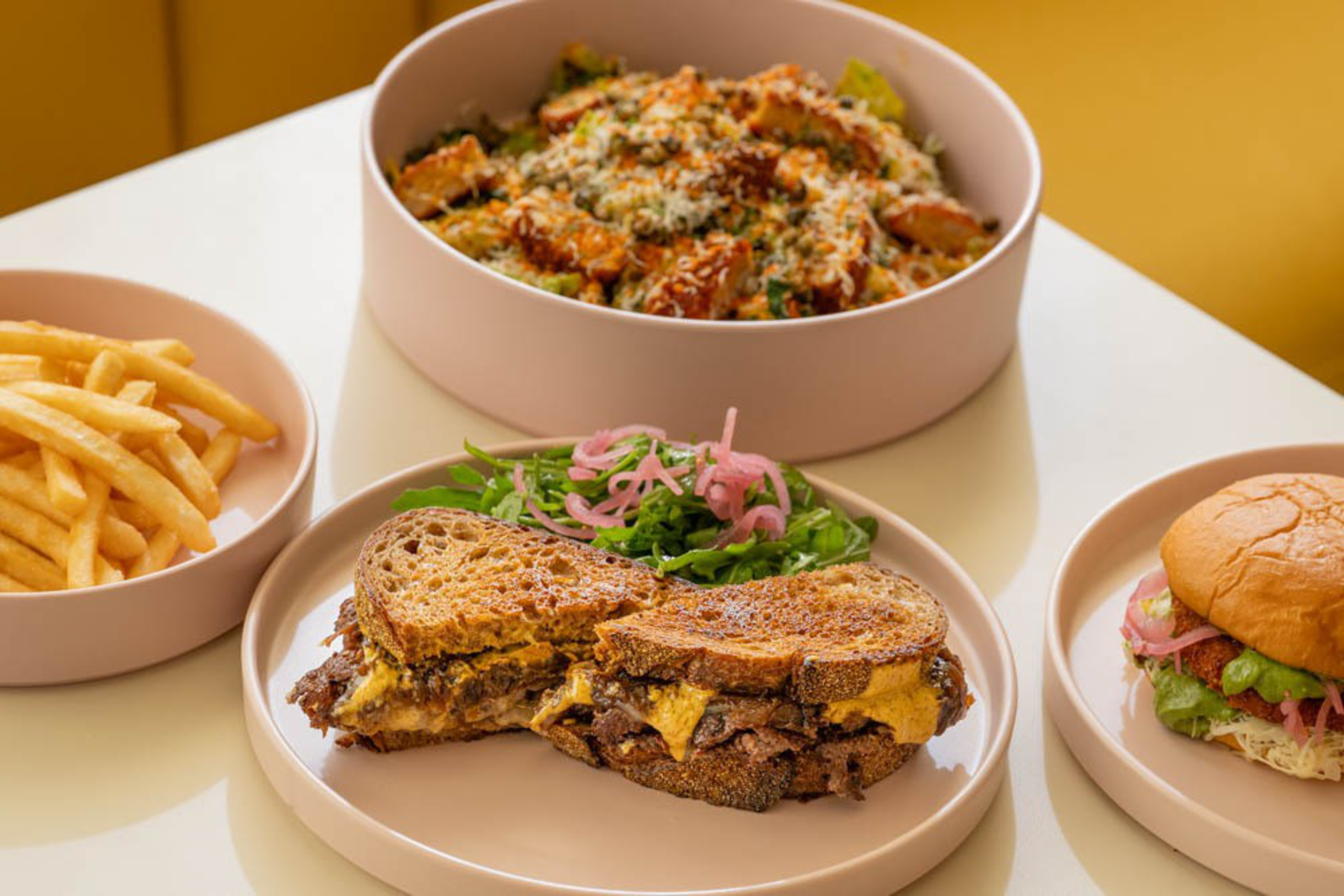 A meal with a grilled cheese sandwich, a side salad with pickled onions, a burger, French fries, and a bowl of pasta topped with cheese and herbs.