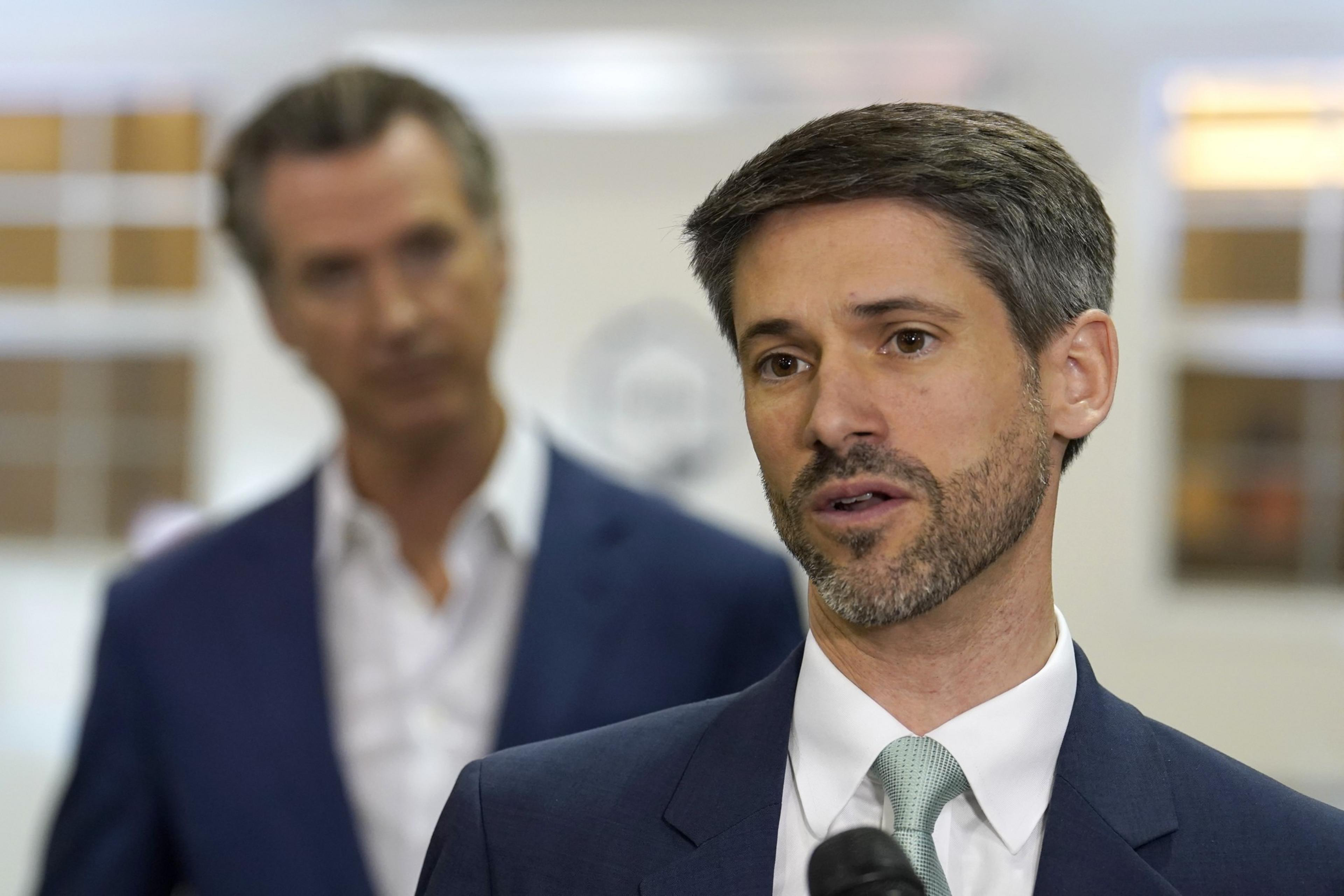 A man with short gray hair, beard, and a suit speaks while a blurred man in a blue suit listens attentively in the background.
