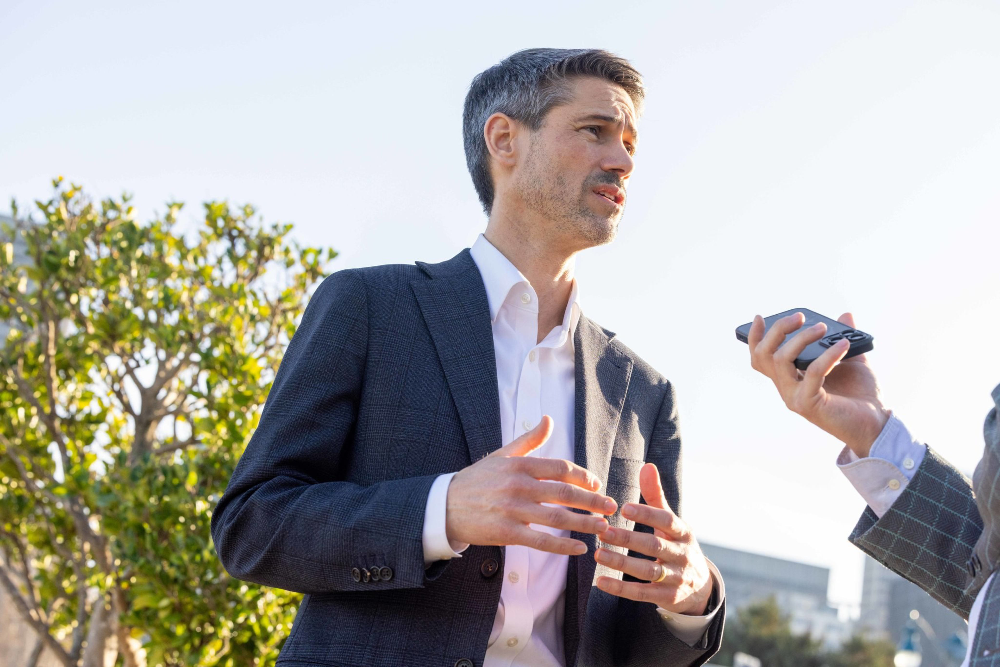 A man in a dark suit and white shirt gestures while speaking to another person holding a voice recorder outdoors on a sunny day.