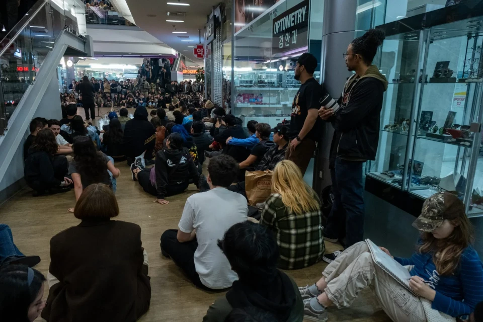 People, sitting on the floor of a mall, listen to a person playing a saxophone in front of a microphone stand. A person in the foreground writes in a notebook.