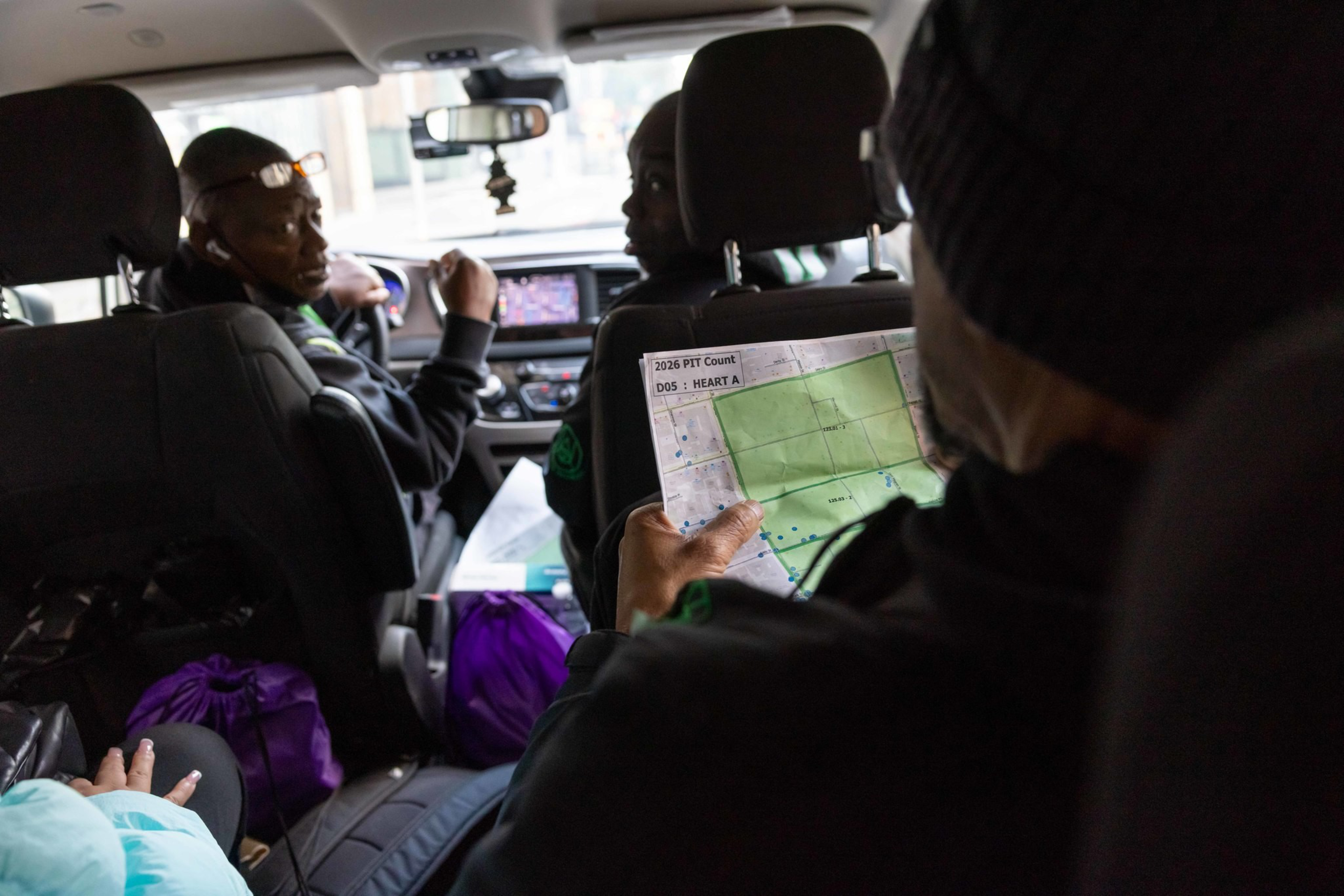 Three people are inside a vehicle, with one holding a map labeled “2026 PIT Count,” while two look forward near the dashboard and purple bags are seen on the floor.