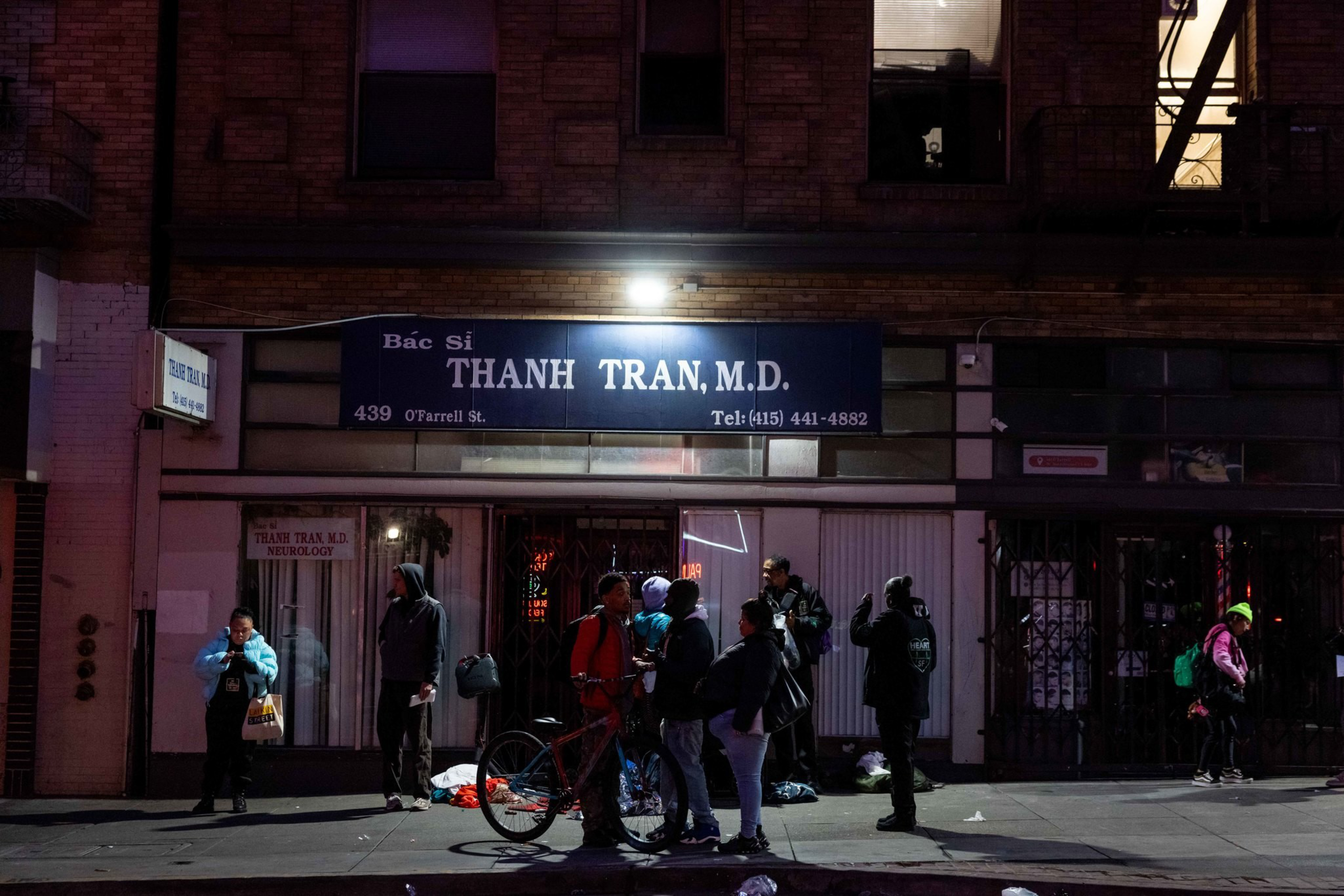 A group of people stand and talk on a dimly lit sidewalk outside a building with a sign for Thanh Tran, M.D. Neurologist at 439 O’Farrell St.