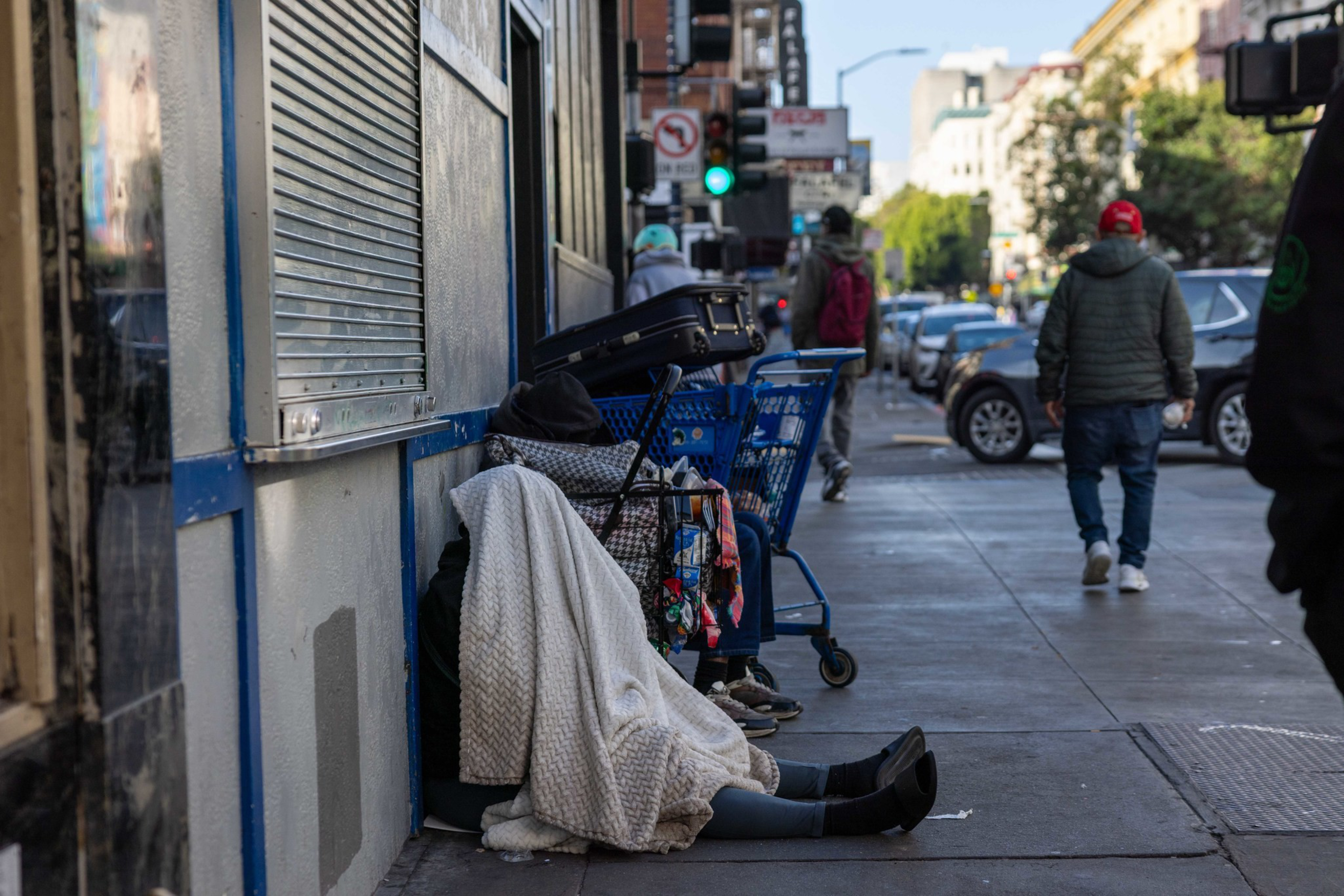A person covered in a blanket sits on a sidewalk leaning against a wall, surrounded by shopping carts and luggage, while pedestrians walk nearby.