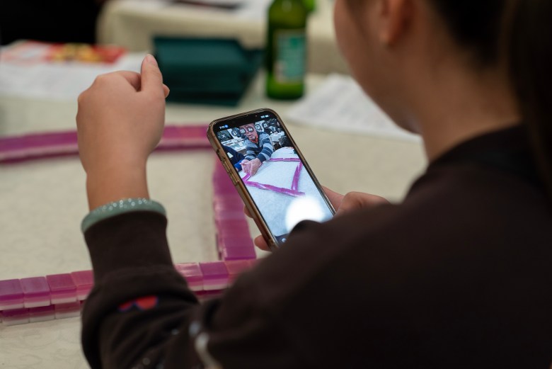 A child sits at a table holding a smartphone, watching a video of an adult demonstrating how to arrange pink plastic counting blocks into a square, with the blocks spread across the tabletop in front of them.