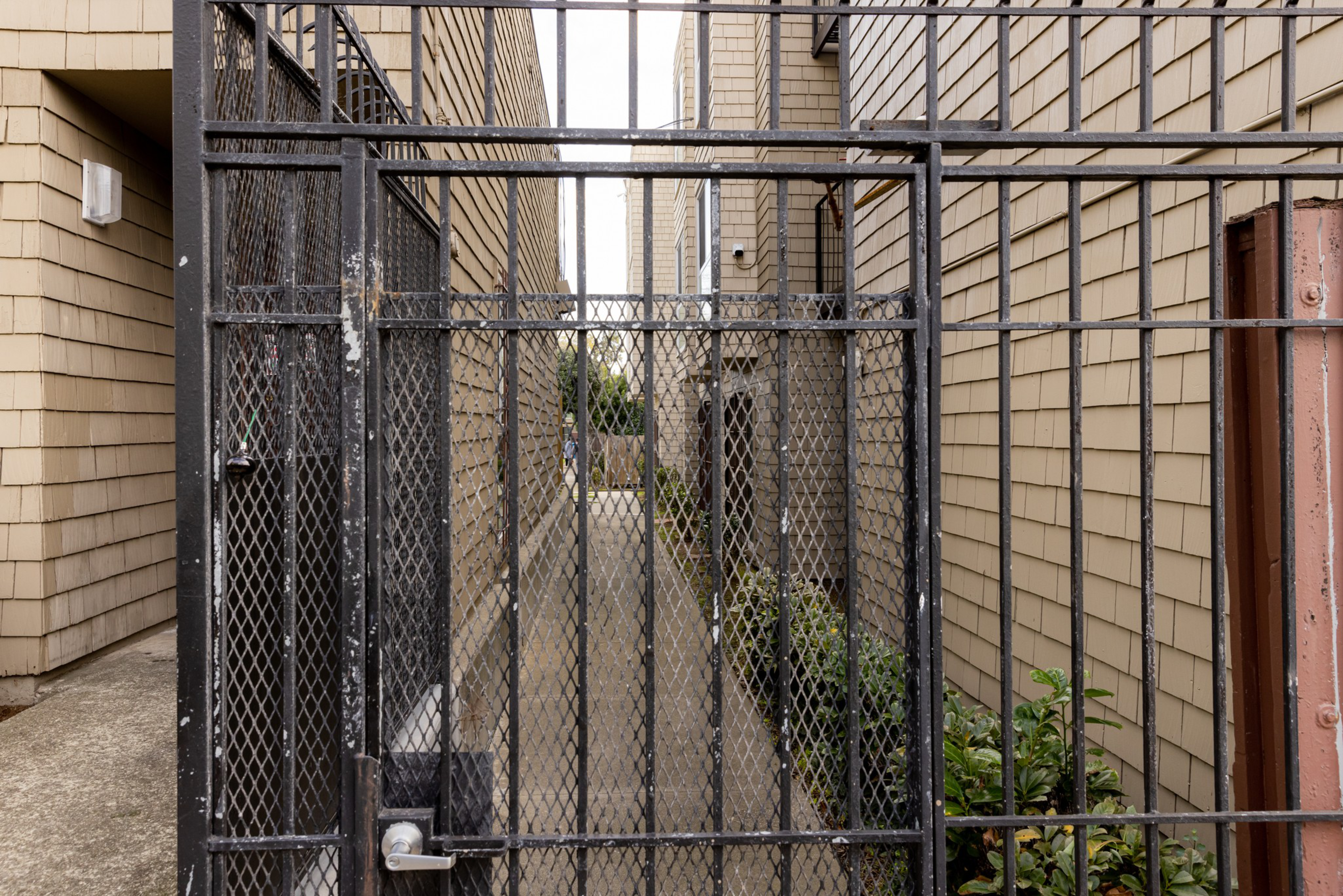 A black metal gate blocks a narrow alleyway between two beige shingled buildings, with some green plants visible along one side.