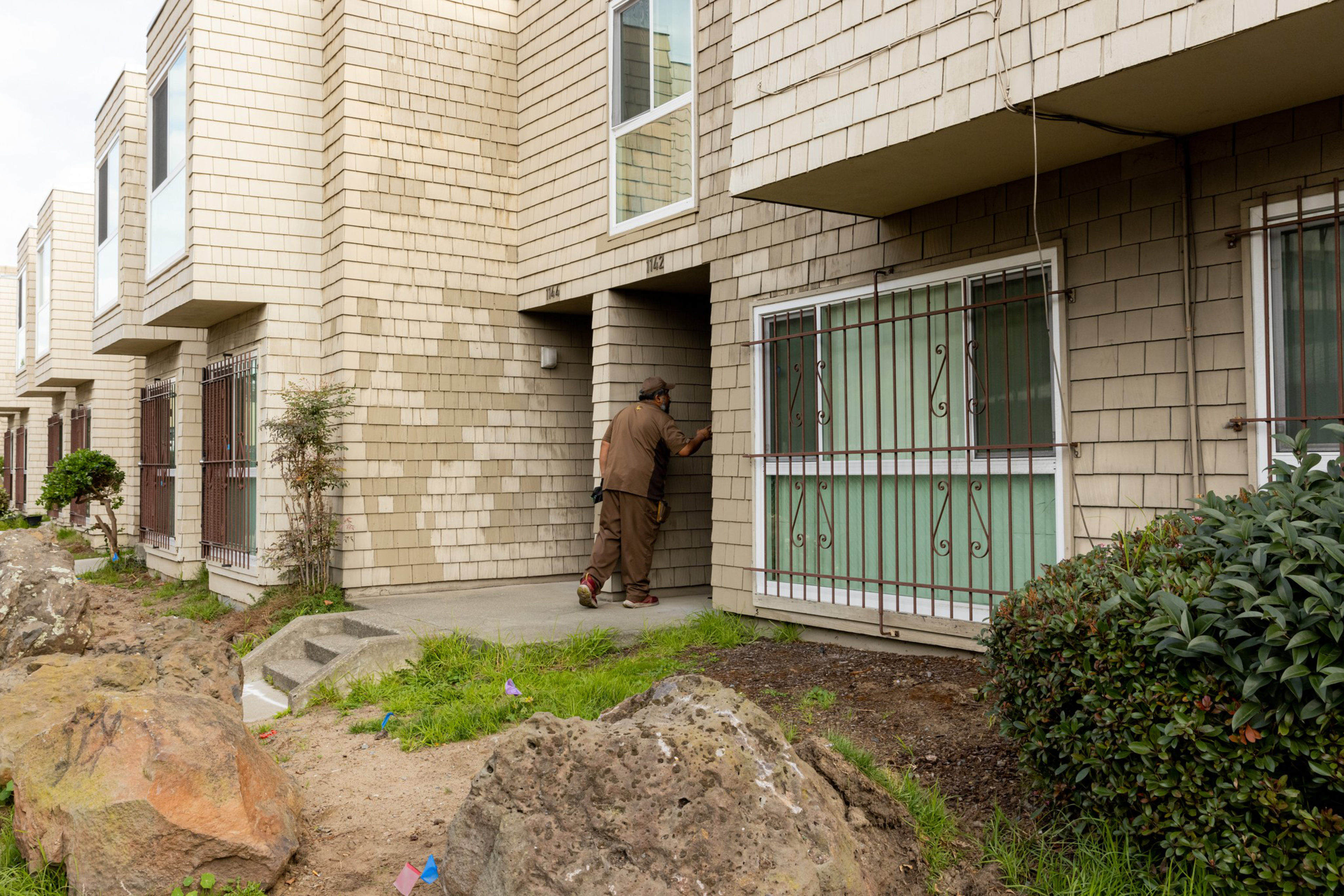 A person dressed in brown is knocking on the door of a beige, multi-unit residential building with barred windows and shrubbery around.