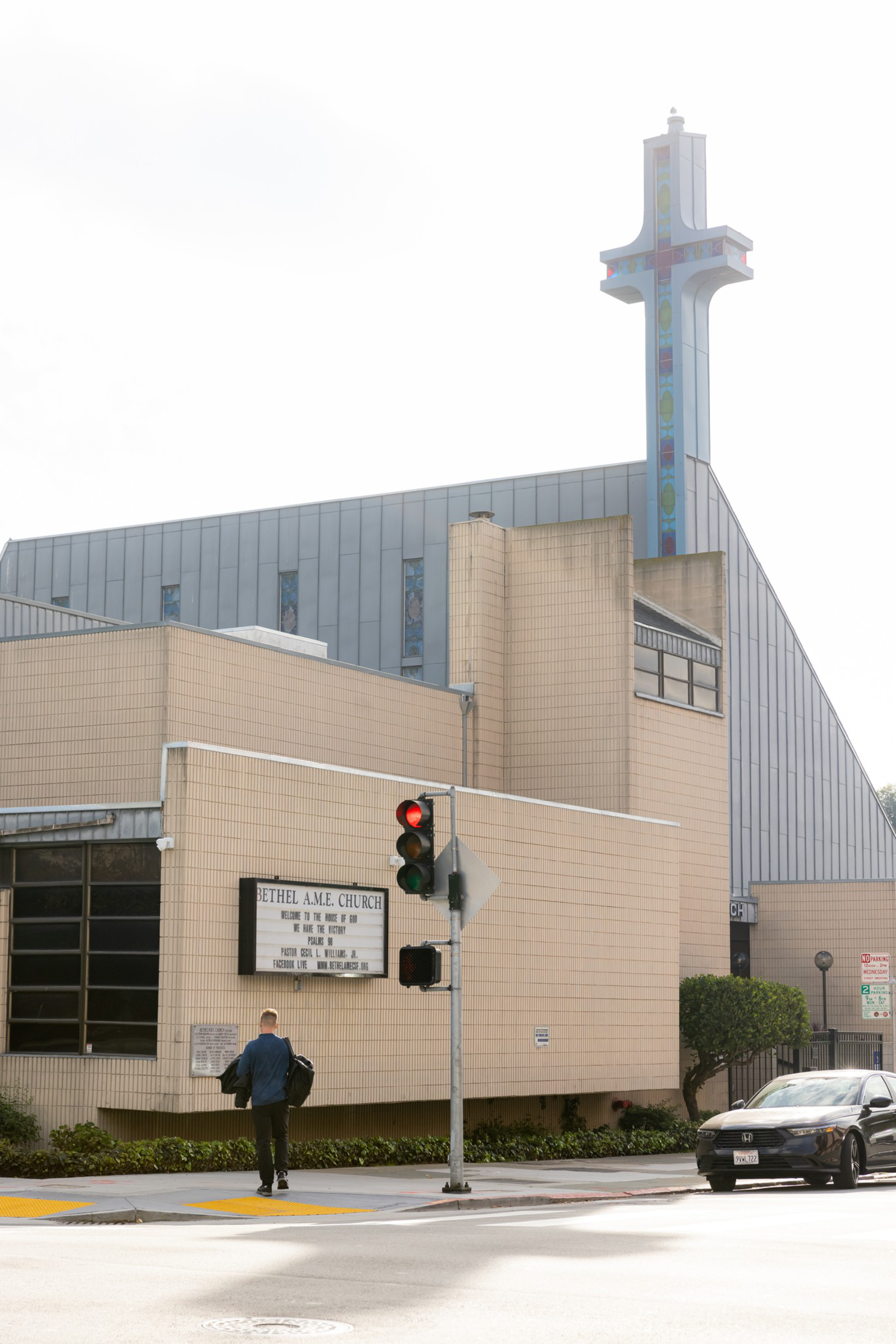 A beige church building with a tall cross, a traffic light showing red, a pedestrian walking, and a car waiting at the corner.