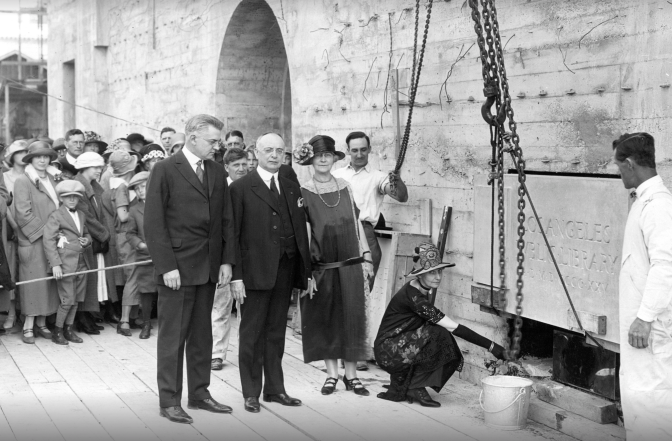 A historical black and white photo from the 1920's, showing two light skinned men and a light skinned woman standing next to a wall. At the bottom of the wall another light skinned woman is crouched down, putting something inside a gap.