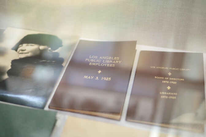 Inside a glass container is a black and white photo of a woman wearing clothes from the 1920's, and two brown books which says Los Angeles Public Library Employees and LAPL Board of Directors