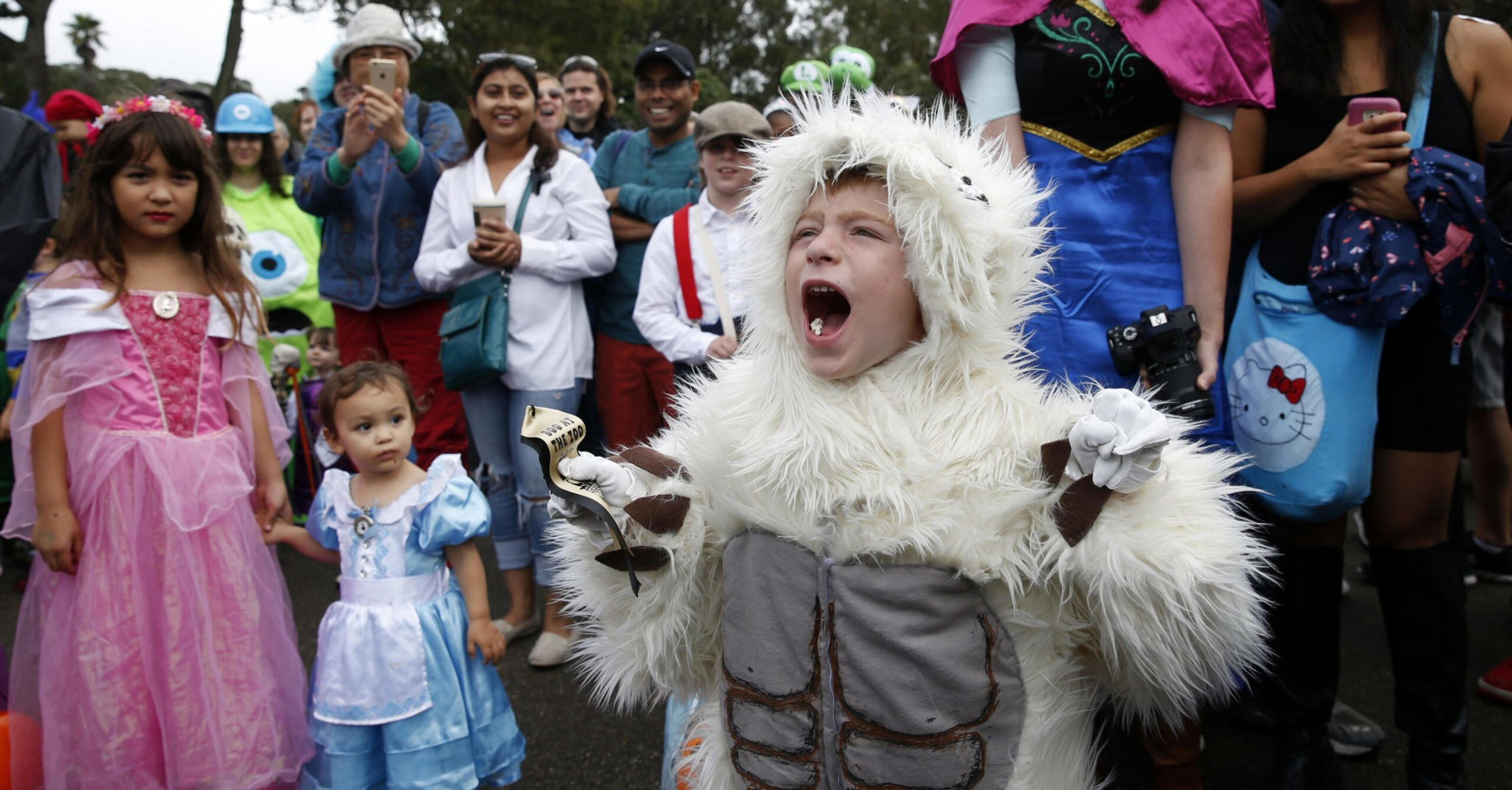 A child in a white furry monster costume roars while other kids dressed as princesses watch, with adults and spectators smiling in the background.