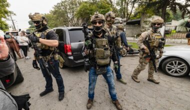 Five immigration agents, dressed in camouflage riot gear and holding weapons, stand in the middle of a street while facing a crowd of bystanders and protesters.