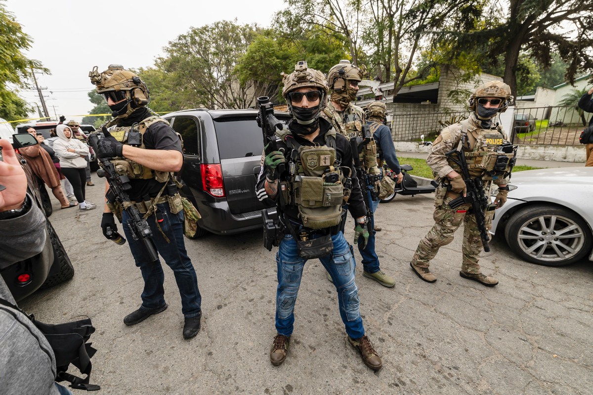 Five immigration agents, dressed in camouflage riot gear and holding weapons, stand in the middle of a street while facing a crowd of bystanders and protesters.