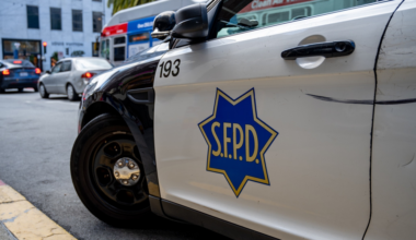A train travels on an elevated track with a busy shipping port in the background. Shipping containers and cranes are visible under a cloudy sky.