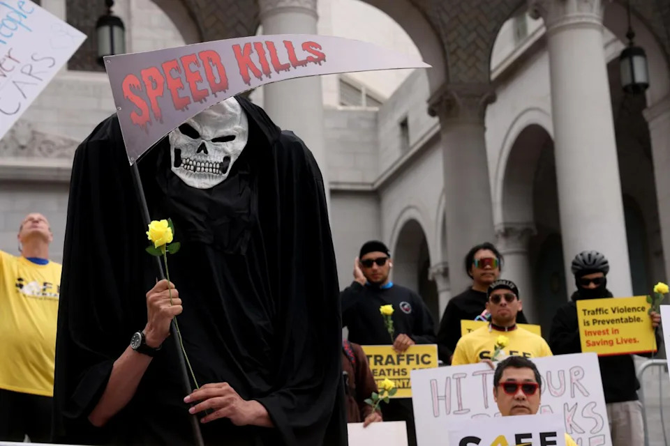 A person in a grim reaper costume holds a scythe emblazoned with the words 'speed kills' in front of other protesters