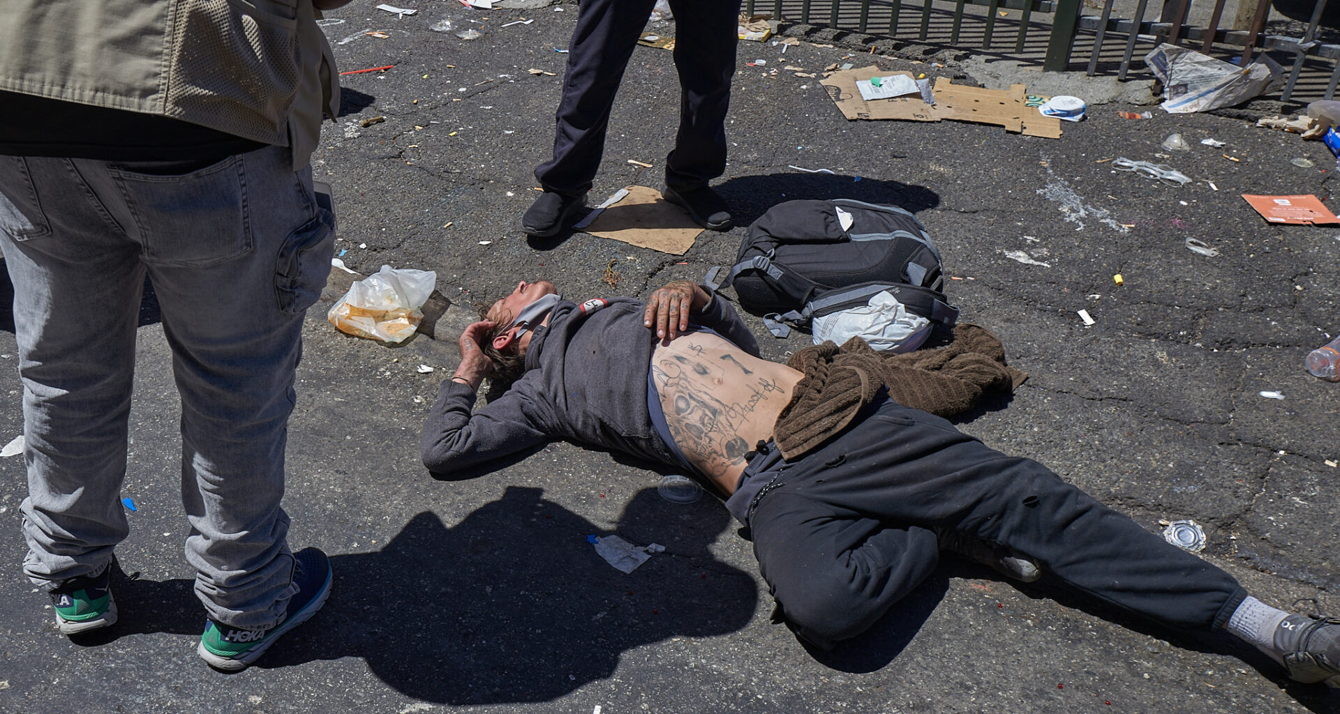 A person lies on a littered street with two people standing nearby. The person on the ground has tattoos on their torso and is surrounded by scattered belongings and debris.