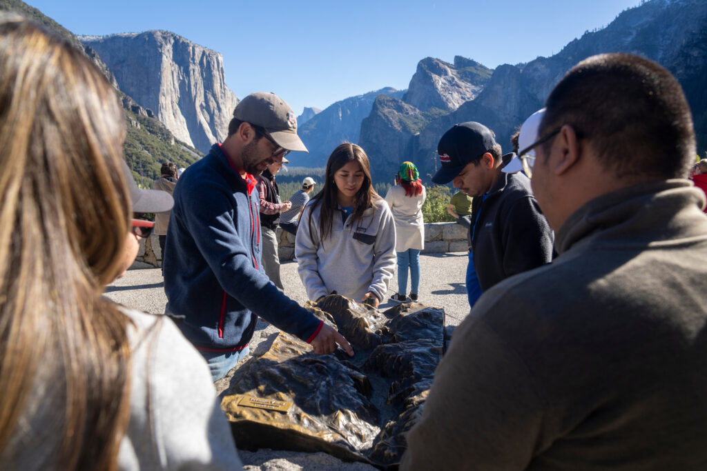 A teacher and his students observe a rock formation with Yosemite Valley in the background