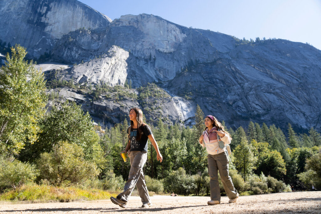Two students hiking on a trail in a forested valley with a large rocky mountain in the background.