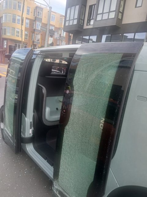 A vehicle with both side doors open shows shattered glass on the windows and ground, suggesting recent damage or vandalism; buildings are visible in the background.