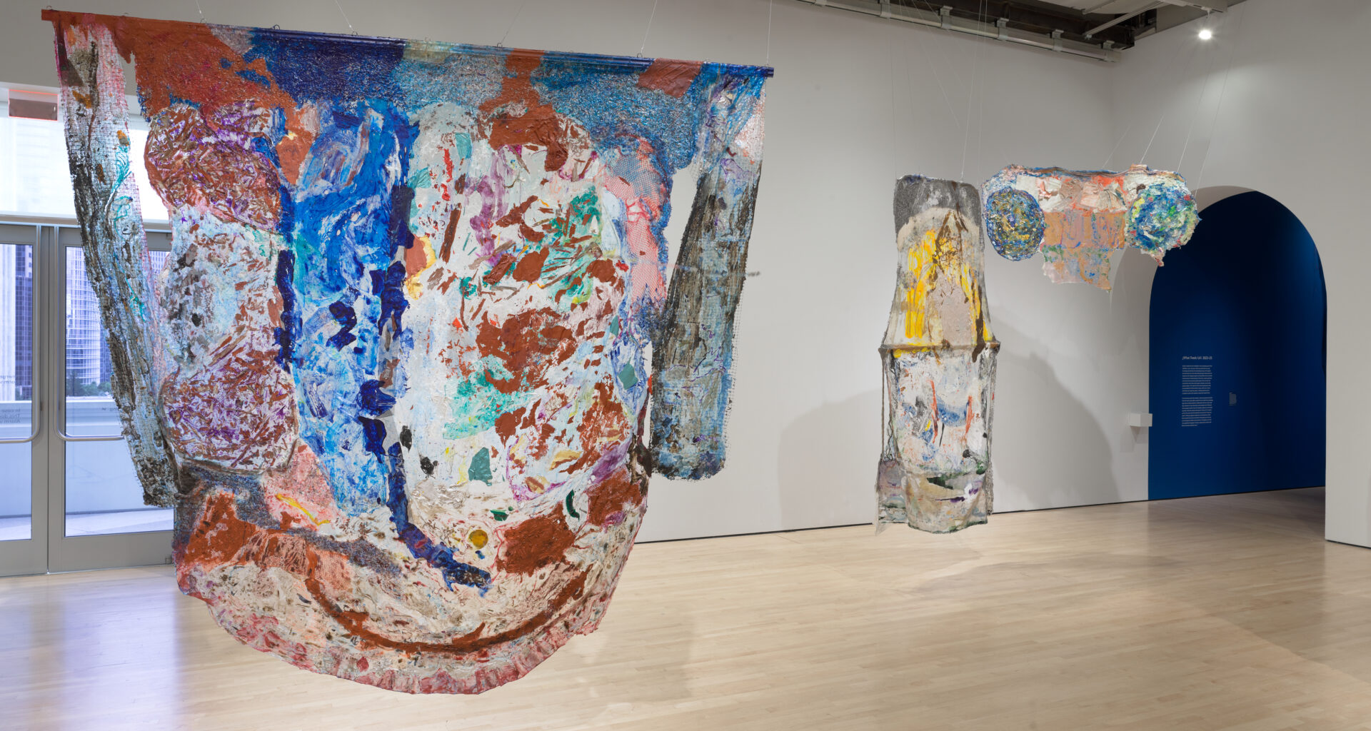 Large, irregularly shaped abstract textile works painted in earthy reds, blues, whites and yellows hang suspended in a spacious museum gallery with light wood floors and white walls.