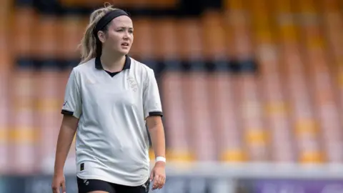 Port Vale Football Club A woman with blonde hair and a black headband is on a football pitch. She wears a white top with gold lettering on the chest that reads "PVFC". The kit is a white T-shirt with a navy collar and navy shorts