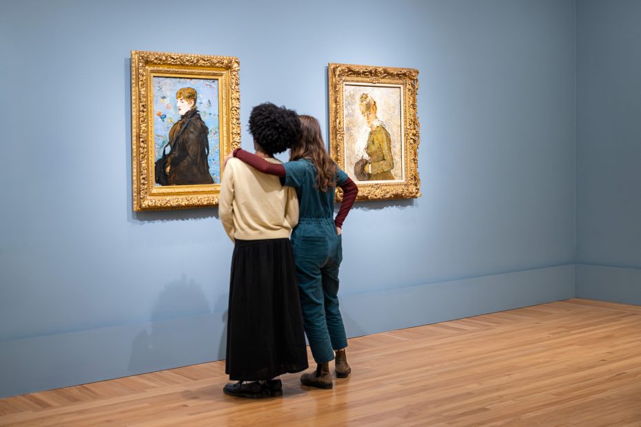 Two people stand arm in arm, facing and observing two framed paintings on a blue wall in an art gallery with wooden floors.