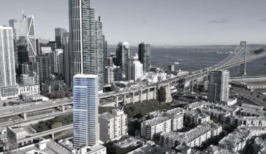A tall modern building highlighted in blue stands among other skyscrapers near a bridge and waterfront in a cityscape.