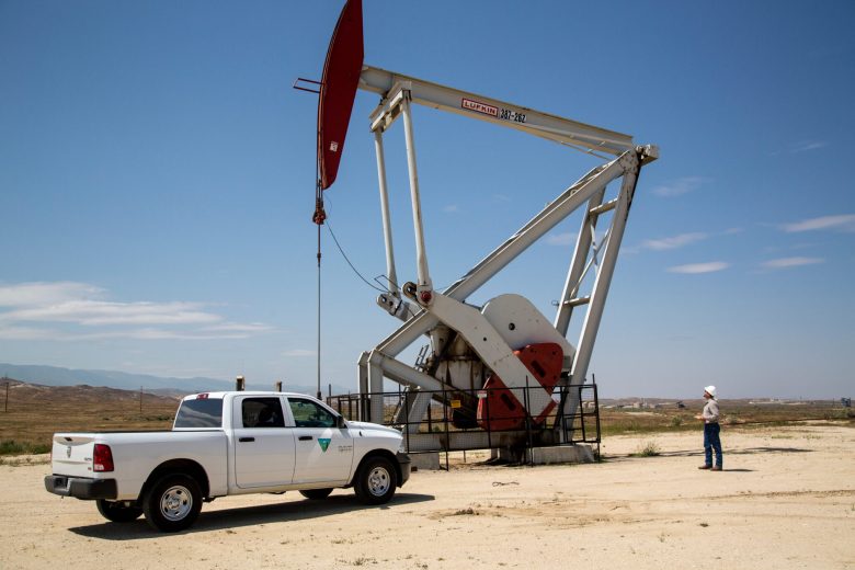 Oil pumpjack in a sandy landscape with a white pickup truck nearby and a person in a hard hat observing the scene.