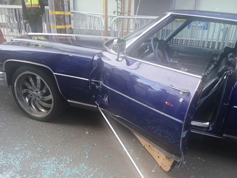 A blue car with a damaged door and shattered glass on the ground; the door is ajar and a metal rod lies beside it. People and barriers are visible in the background.