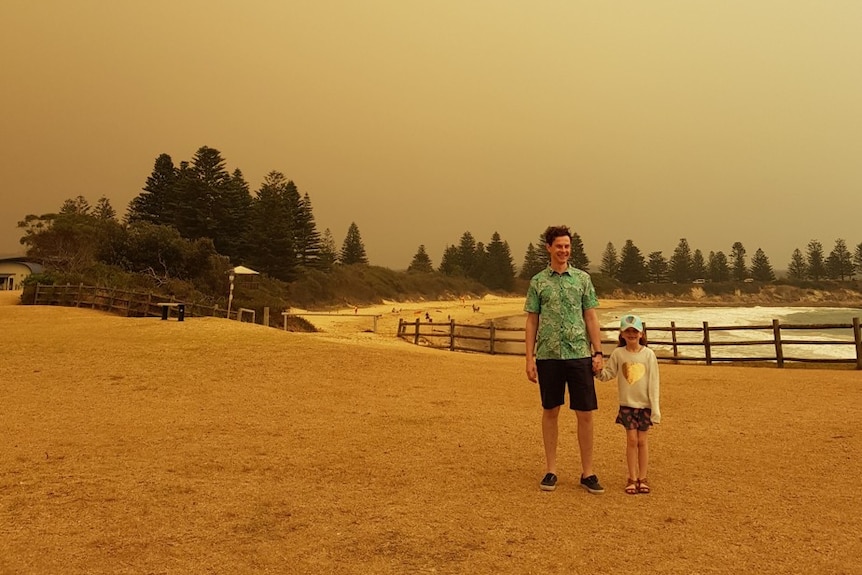 A man and a child standing at the beach while a bushfire burns behind them. 
