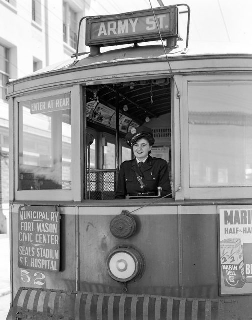 A woman in uniform is seated at the controls of a streetcar marked "Army St." with route signs for various San Francisco locations visible on the front.