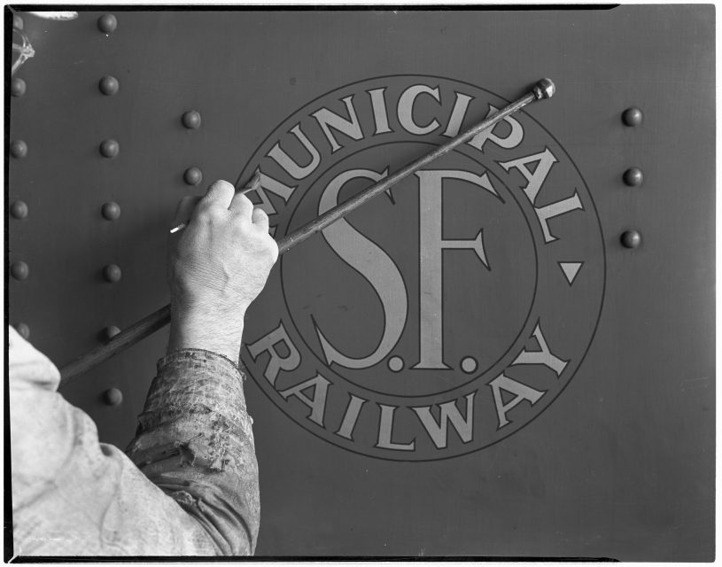 A person paints or touches up the "S.F. Municipal Railway" emblem on a metal surface.
