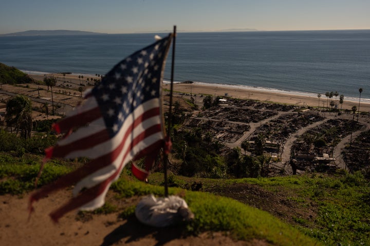 A tattered U.S. flag flaps in the wind over the remains of a mobile home park that was destroyed a year ago in the Palisades Fire in the Pacific Palisades neighborhood of Los Angeles.