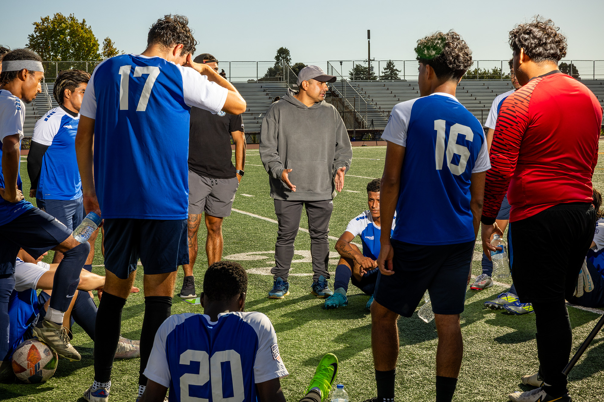 Soccer players in blue jerseys huddle around their coach, who wears gray sweats.