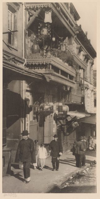 Black-and-white photo of people walking down a narrow street lined with multi-story buildings featuring ornate balconies and hanging lanterns.