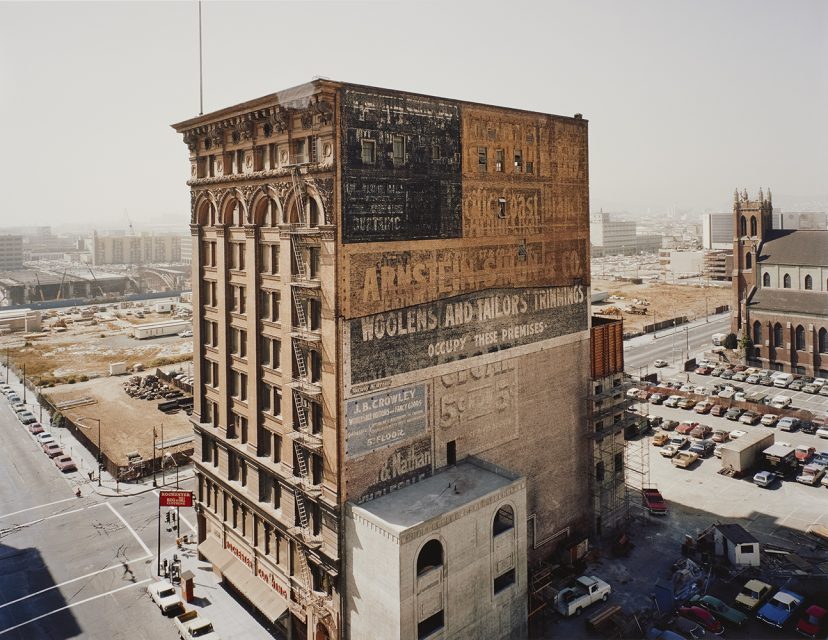 A tall brick building with faded painted advertisements stands at a city intersection, surrounded by parking lots, construction sites, and distant cityscape views.