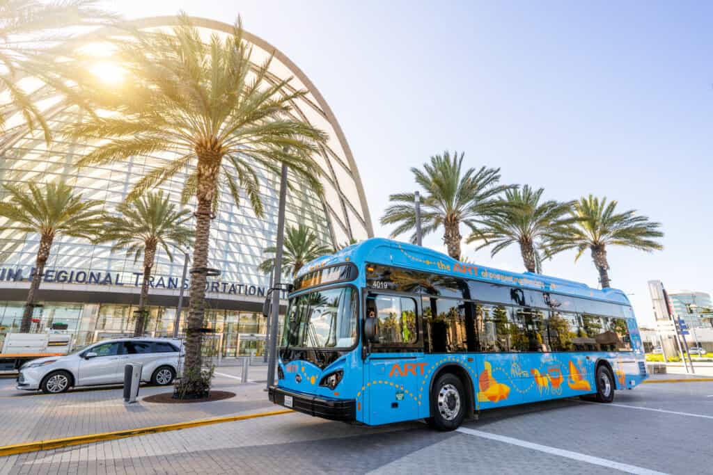 A vivid blue shuttle bus rolls past palm trees outside the glassy entrance of Universal CityWalk on a sun-filled day. Disneyland transportation system shutting down