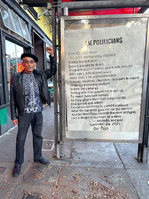A man in a black hat and jacket stands next to a large sign titled "THE POLITICIANS," which has a poem about war, casualties, and media, displayed on a city sidewalk.
