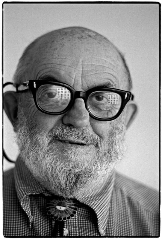 Black and white portrait of an older man with a bald head, bushy white beard, thick-rimmed glasses, and a patterned shirt. The reflection of windows appears in his glasses.