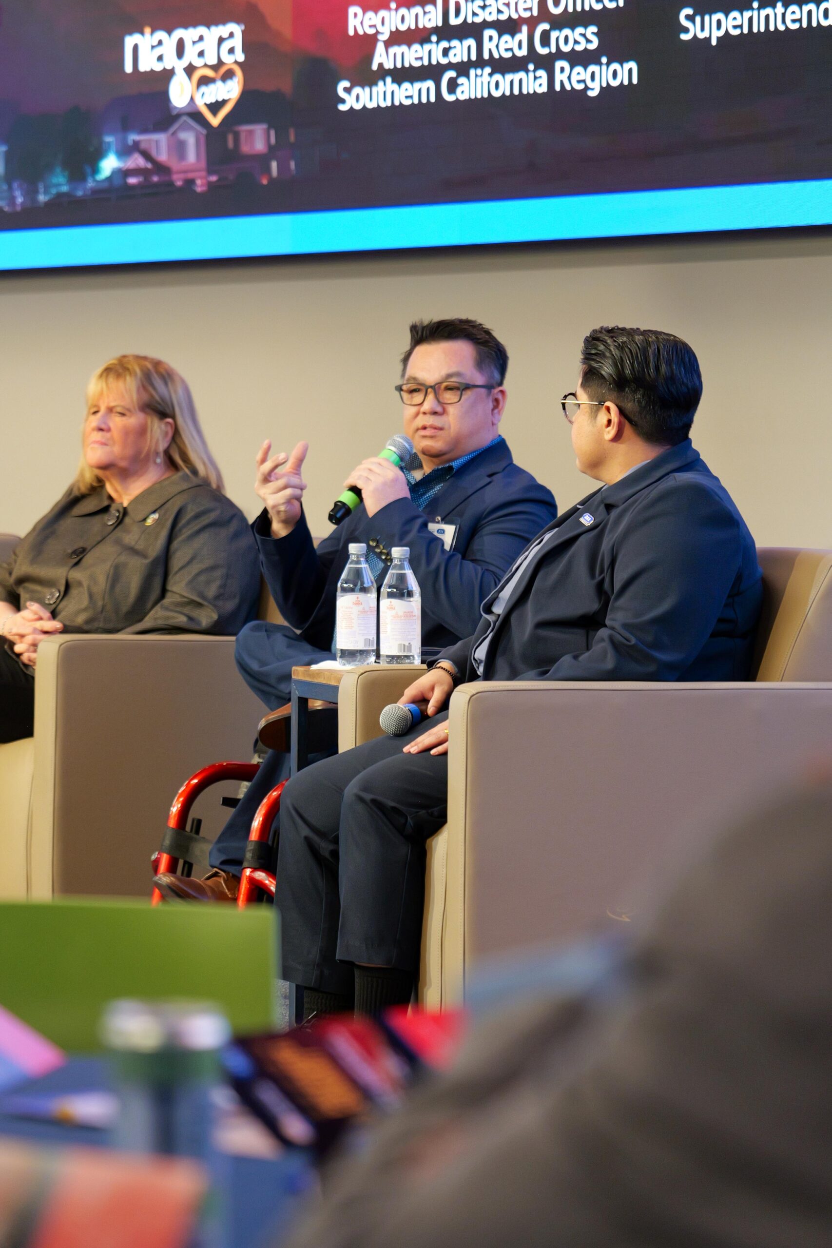 Dr. Stefan Bean speaks into a microphone during a panel discussion at the Be Prepared OC Summit, seated between Debbie Leahy of the American Red Cross on the left and Amy Arambulo of 211 Orange County on the right.