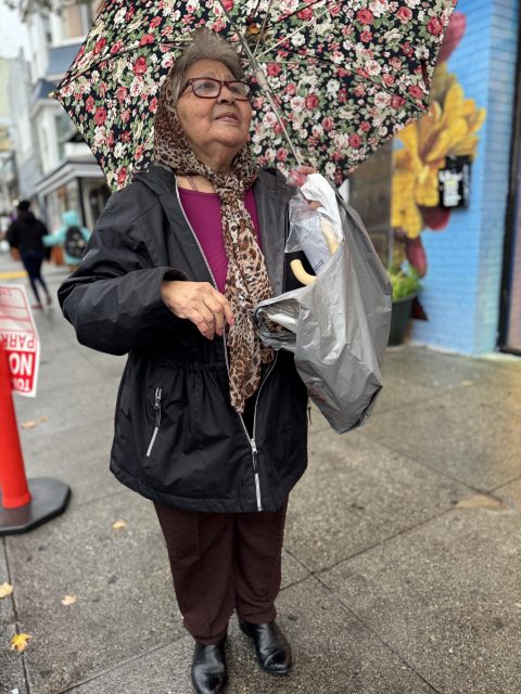 An older woman stands on a city sidewalk holding a floral umbrella and a shopping bag, wearing a scarf and glasses, with a colorful mural in the background.