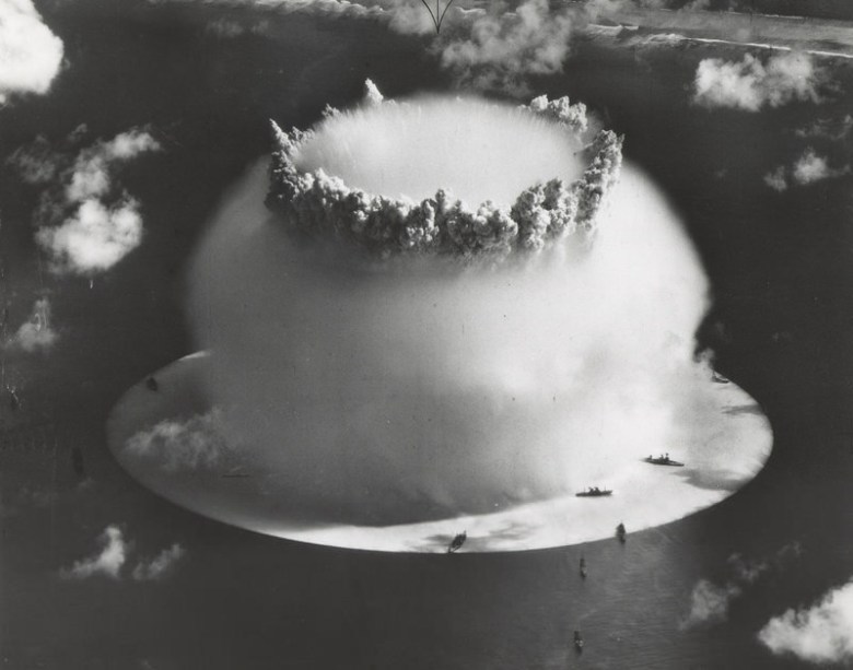 Aerial black and white image of a mushroom cloud over the ocean with ships close to the blast