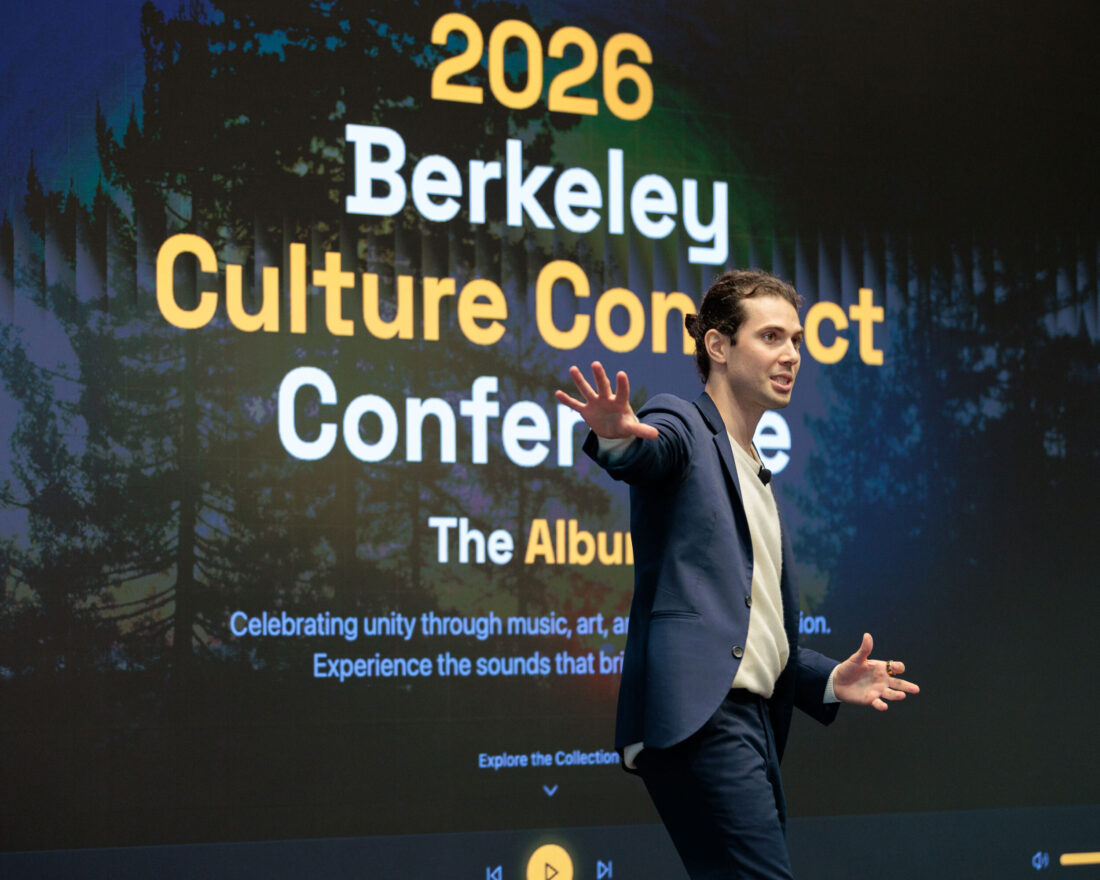 A young man in a suit and white dress shirt stretches his arm out in front of a screen that reads "2026 Berkeley Culture Connect Conference - The Album"