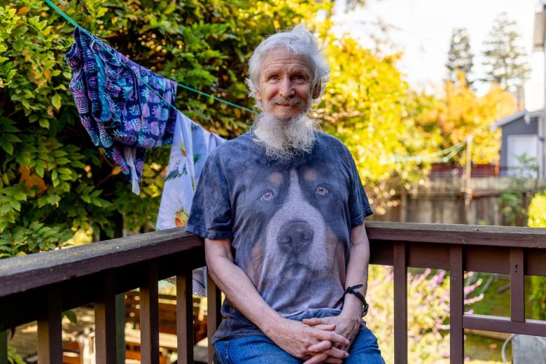 A man with gray hair and a gray beard, wearing a t-shirt with an illustration of a hound's face, sits smiling with his hands folded on a backyard deck next to a clothesline with green trees in the background.
