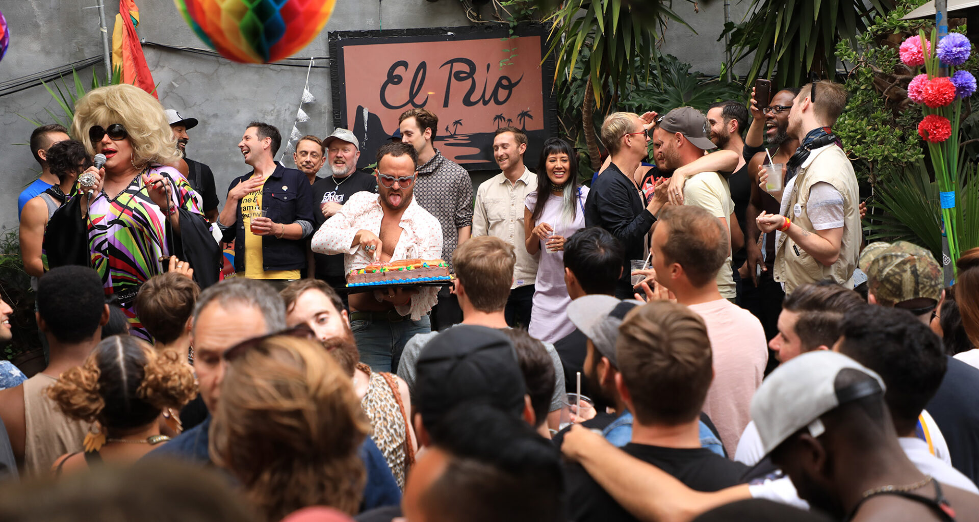Crowd mingling on a bar's outdoor patio. A pink sign in the back reads, "El Rio."