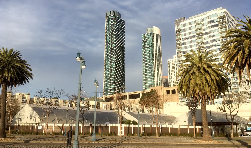 Towers rise behind low huts that form a speclialized homeless shelter called a Navigation Center in San Francisco. 