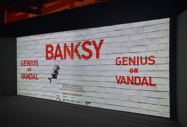 Spraypainted brick wall advertising Banksy: Genius or Vandal exhibition